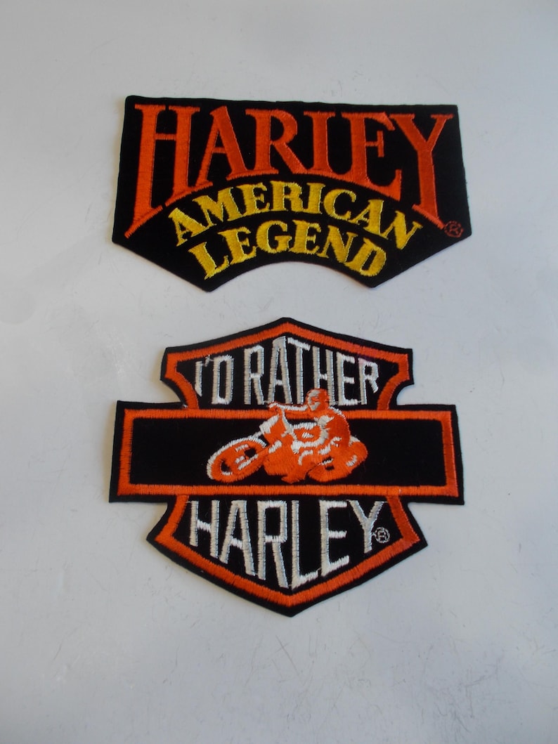 May include: Two Harley-Davidson patches. The top patch reads "HARLEY AMERICAN LEGEND" in orange and yellow lettering on a black background. The bottom patch features a motorcycle graphic and the word "HARLEY".