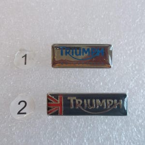 May include: Two silver metal pins with the word "TRIUMPH" in blue lettering. The first pin has a silver background. The second pin has a black background with a red, white, and blue Union Jack flag in the upper left corner.