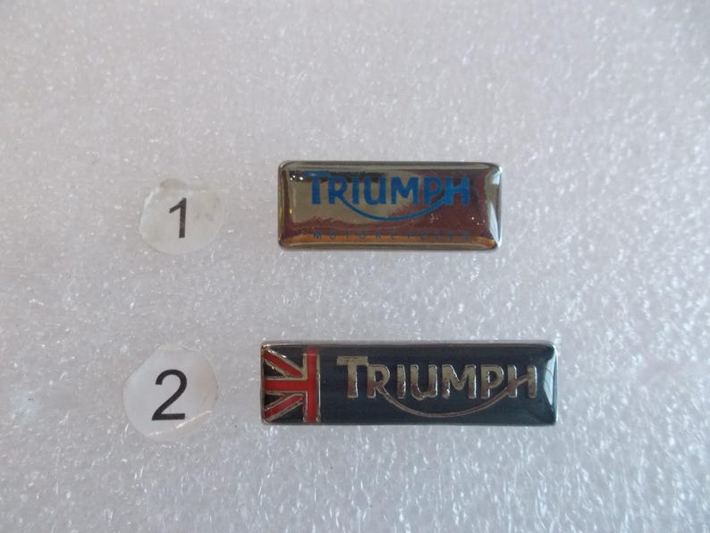 May include: Two silver metal pins with the word "TRIUMPH" in blue lettering. The first pin has a silver background. The second pin has a black background with a red, white, and blue Union Jack flag in the upper left corner.