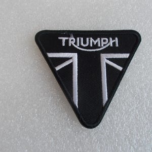 K&ouml;nnte beinhalten: Schwarzer Triumph-Aufn&auml;her in Dreiecksform. Der Aufn&auml;her zeigt das Wort "TRIUMPH" in Wei&szlig; &uuml;ber einem wei&szlig;en Design, das an den Union Jack erinnert. Ideal f&uuml;r Motorrad-Enthusiasten.
