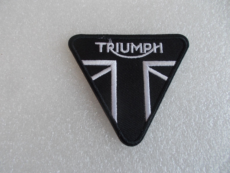 K&ouml;nnte beinhalten: Schwarzer Triumph-Aufn&auml;her in Dreiecksform. Der Aufn&auml;her zeigt das Wort "TRIUMPH" in Wei&szlig; &uuml;ber einem wei&szlig;en Design, das an den Union Jack erinnert. Ideal f&uuml;r Motorrad-Enthusiasten.