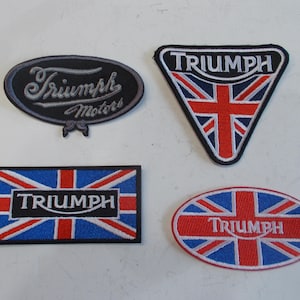 May include: Four embroidered Triumph patches in various shapes: oval, rectangular, and triangular. Each patch features the Triumph logo and Union Jack flag design in red, white, and blue. The oval patch also includes the words "Triumph Motors".