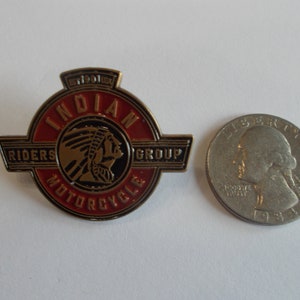 May include: A circular metal pin featuring the Indian Motorcycle Riders Group logo with a red and black enamel finish. The design includes a profile of a person wearing a headdress, the text EST 1901 USA, and is shown next to a US quarter coin.
