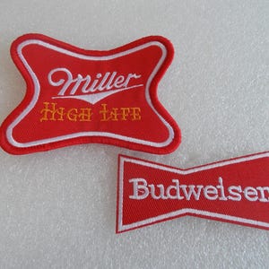 May include: Two embroidered patches with red backgrounds and white borders. The top patch is shaped like a bow tie and features the white text Miller and yellow text High Life. The bottom patch is a red trapezoid with the white text Budweiser.