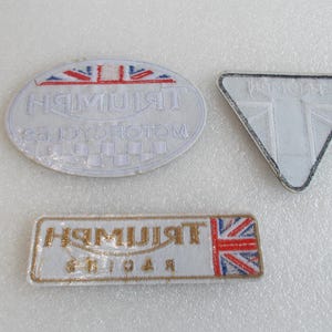K&ouml;nnte beinhalten: Drei wei&szlig;e, gestickte Aufn&auml;her mit dem Triumph-Logo. Einer ist oval, einer rechteckig und einer dreieckig. Der ovale Aufn&auml;her hat ein Schachbrettmuster.