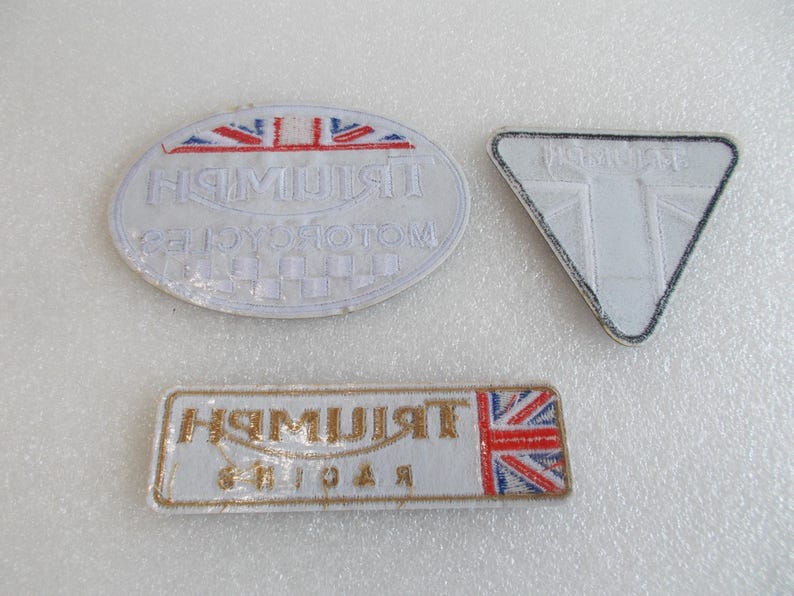 K&ouml;nnte beinhalten: Drei wei&szlig;e, gestickte Aufn&auml;her mit dem Triumph-Logo. Einer ist oval, einer rechteckig und einer dreieckig. Der ovale Aufn&auml;her hat ein Schachbrettmuster.