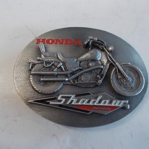May include: Oval-shaped, silver-colored belt buckle featuring a detailed motorcycle design. The word "HONDA" is in red at the top, and "Shadow" is in a red and white design at the bottom. The motorcycle is a detailed, three-dimensional rendering.