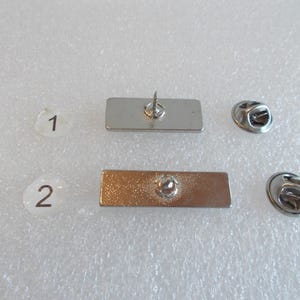 May include: Two silver metal rectangular pins with a safety pin closure. The pins are blank and have a smooth surface.