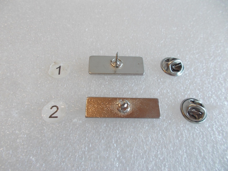 May include: Two silver metal rectangular pins with a safety pin closure. The pins are blank and have a smooth surface.