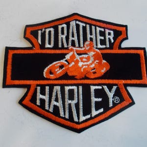 May include: Embroidered patch with a black background and orange trim. The patch features the text "I'D RATHER HARLEY" in white, with an orange motorcycle and rider graphic in the center. The patch is in a shield shape.