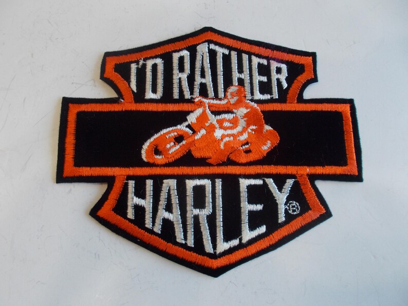 May include: Embroidered patch with a black background and orange trim. The patch features the text "I'D RATHER HARLEY" in white, with an orange motorcycle and rider graphic in the center. The patch is in a shield shape.