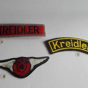 May include: Three embroidered patches with the name "Kreidler" on them. One patch is red with black lettering and a gray border. Another patch is black with yellow lettering and a curved shape. The third patch is red with a white winged design and a black circle with a white cross.