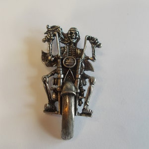 May include: A silver-toned metal pin of a skeleton riding a motorcycle. The skeleton is wearing a leather jacket and has a skull for a head. The motorcycle has a single front wheel and a detailed design.