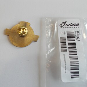 May include: A gold-toned metal lapel pin with a circular center and two side extensions, featuring a butterfly clutch backing. It is placed next to a clear plastic bag with a white label reading Indian Motorcycle, 2869832, and IMRG LAPEL PIN.