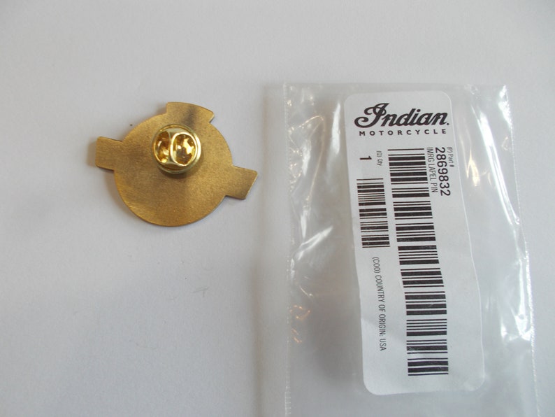 May include: A gold-toned metal lapel pin with a circular center and two side extensions, featuring a butterfly clutch backing. It is placed next to a clear plastic bag with a white label reading Indian Motorcycle, 2869832, and IMRG LAPEL PIN.