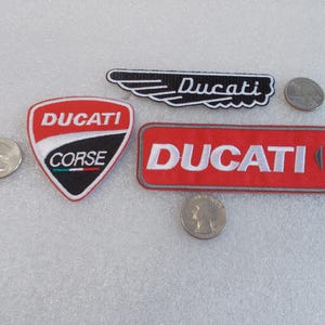 May include: Three embroidered patches featuring the Ducati brand. One is a shield-shaped Ducati Corse patch, one is a rectangular red Ducati patch, and one is a black winged Ducati logo. Three US quarters are placed nearby for scale reference.