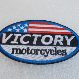 May include: Oval patch with the text "VICTORY motorcycles". The top of the patch features the American flag with red and white stripes and white stars on a blue background. The patch has a blue border.