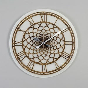 May include: A circular wall clock with a white face and a decorative wooden frame. The frame has Roman numerals and an intricate floral design in the centre. The clock hands are silver, and the clock is mounted on a grey wall.