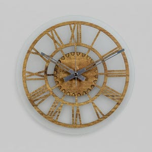 Puede incluir: Un reloj de pared redondo con un marco de madera dorada y números romanos. Las manecillas del reloj son plateadas y el fondo es blanco esmerilado. El diseño del reloj es abierto, con los mecanismos internos visibles.