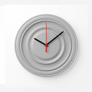 May include: A circular wall clock with a light grey face featuring concentric circular ridges. The clock has two black hour and minute hands and a thin, straight red second hand extending vertically from the centre point of the clock face.