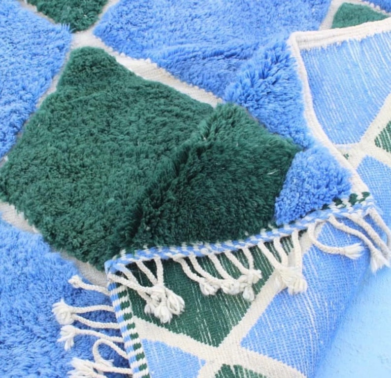 May include: A blue and green geometric patterned rug with a fringe border. The rug is made of soft, fluffy fibers.