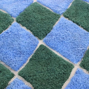 May include: A close-up of a blue and green shag rug with a diamond pattern. The rug is made of soft, fluffy fibers and has a white background.