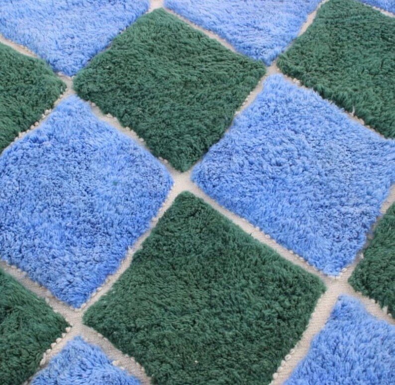 May include: A close-up of a blue and green shag rug with a diamond pattern. The rug is made of soft, fluffy fibers and has a white background.