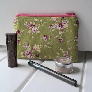 May include: A green floral print makeup bag with a pink zip, alongside a brown lipstick tube, a black eyeliner, and a small eyeshadow pot. The bag displays a pattern of purple and yellow flowers.