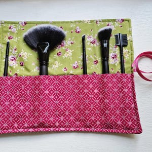 May include: A fabric makeup brush roll featuring a green floral interior and a magenta exterior with a white geometric circle pattern. Five black makeup brushes are tucked into the pockets, and a magenta ribbon is attached to the side.