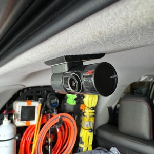 May include: A black dashcam mounted to the ceiling of a car. The camera has a lens on the front and a small red light on the side.