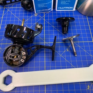 May include: Disassembled black fishing reel components, including a reel body, handle, and various internal parts, arranged on a blue grid surface. A light gray wrench is also present.