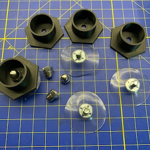 May include: A set of four black plastic suction cup mounts with silver metal screws. The mounts are on a blue grid surface. There are four silver metal nuts on the surface.