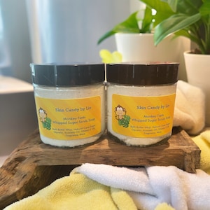 May include: Two glass jars with black lids containing a whipped sugar scrub soap. The jars have a yellow label with a cartoon monkey and the text "Skin Candy by Liv" and "Monkey Farts Whipped Sugar Scrub Soap".