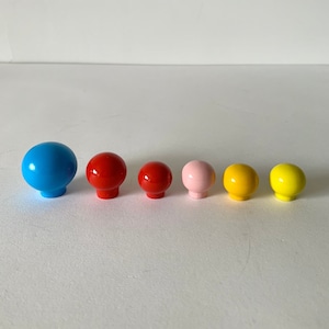 Könnte beinhalten: Sechs bunte, runde Knöpfe in einer Reihe. Die Knöpfe sind in Blau, Rot, Rosa und Gelb gehalten. Die Knöpfe haben eine glänzende Oberfläche und eine pilzartige Form. Wahrscheinlich sind es Schrank- oder Schubladengriffe.