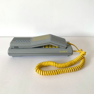 May include: A gray vintage rotary phone with a yellow coiled cord. The phone has a flip-up handset and the word "swatch" printed on the front.