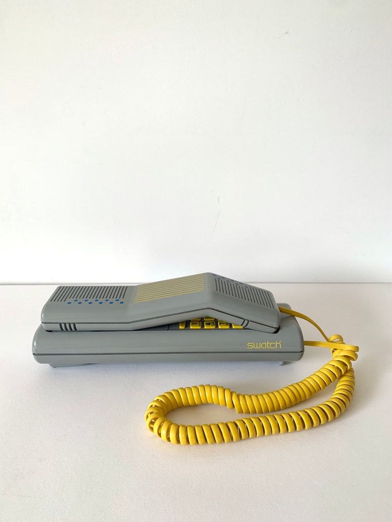 May include: A gray vintage rotary phone with a yellow coiled cord. The phone has a flip-up handset and the word "swatch" printed on the front.