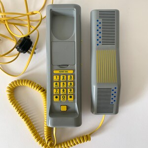 May include: A gray rotary phone with a yellow coiled cord and a separate handset. The phone has a keypad with numbers and letters, and a small display screen that says "NAME DIAL".