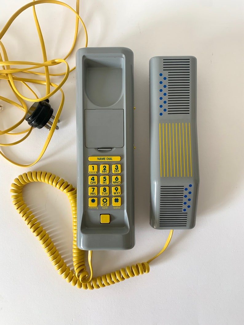 May include: A gray rotary phone with a yellow coiled cord and a separate handset. The phone has a keypad with numbers and letters, and a small display screen that says "NAME DIAL".