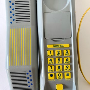 May include: A gray corded phone with a yellow cord and a gray and yellow keypad. The keypad has the letters ABC, DEF, GHI, JKL, MNO, PRS, TUV, WXY, QZ, and the symbols * and #. The phone has a gray and yellow name dial button, a gray and yellow memory button, a gray and yellow oper button, and a gray and yellow redial button.