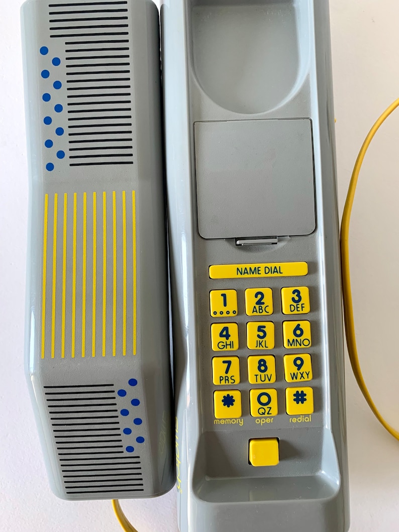 May include: A gray corded phone with a yellow cord and a gray and yellow keypad. The keypad has the letters ABC, DEF, GHI, JKL, MNO, PRS, TUV, WXY, QZ, and the symbols * and #. The phone has a gray and yellow name dial button, a gray and yellow memory button, a gray and yellow oper button, and a gray and yellow redial button.