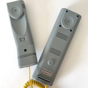 May include: Two gray vintage-style corded phones with a yellow coiled cord. The phones are labeled "Swatch Twin Phone".