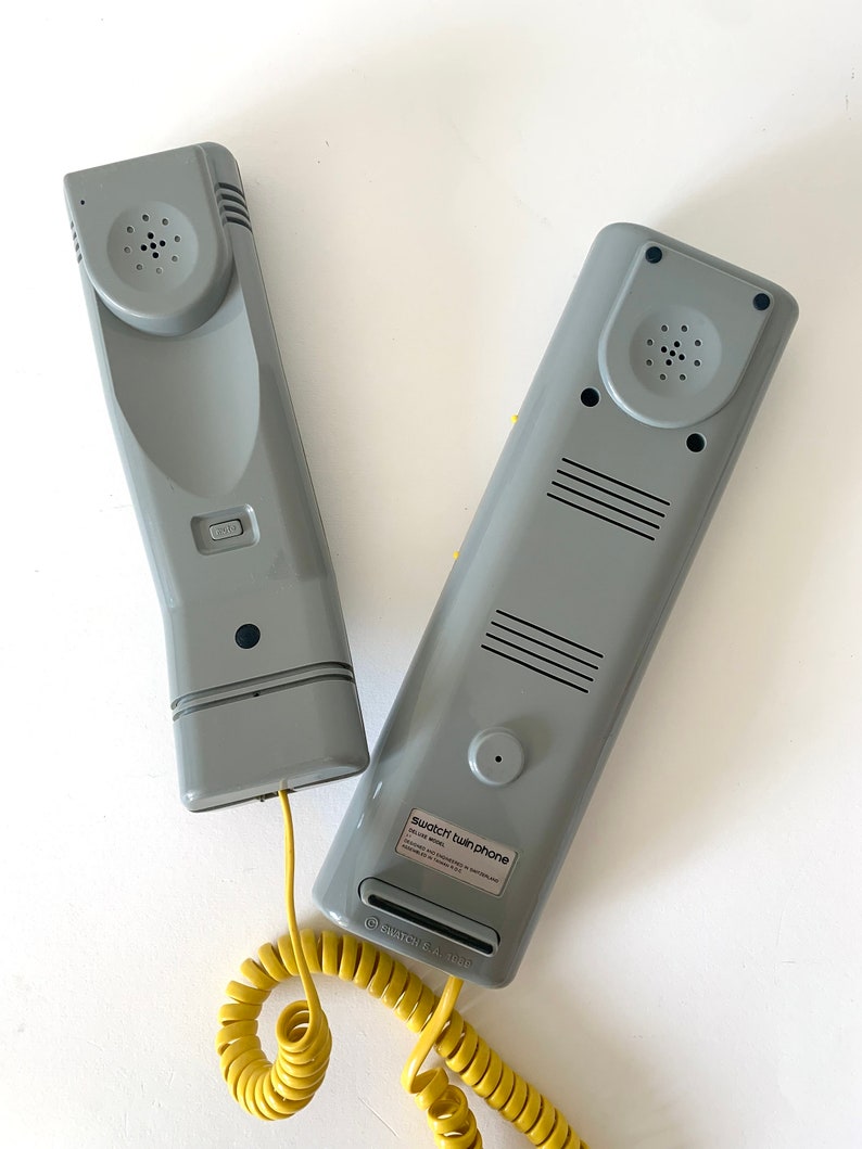 May include: Two gray vintage-style corded phones with a yellow coiled cord. The phones are labeled "Swatch Twin Phone".