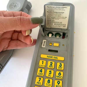 May include: A gray phone with a yellow keypad and a small gray flip-up cover. The cover has instructions for programming the phone in Italian. The keypad has numbers 1 through 9, a star, a circle with the letter "O" inside, and a pound sign.