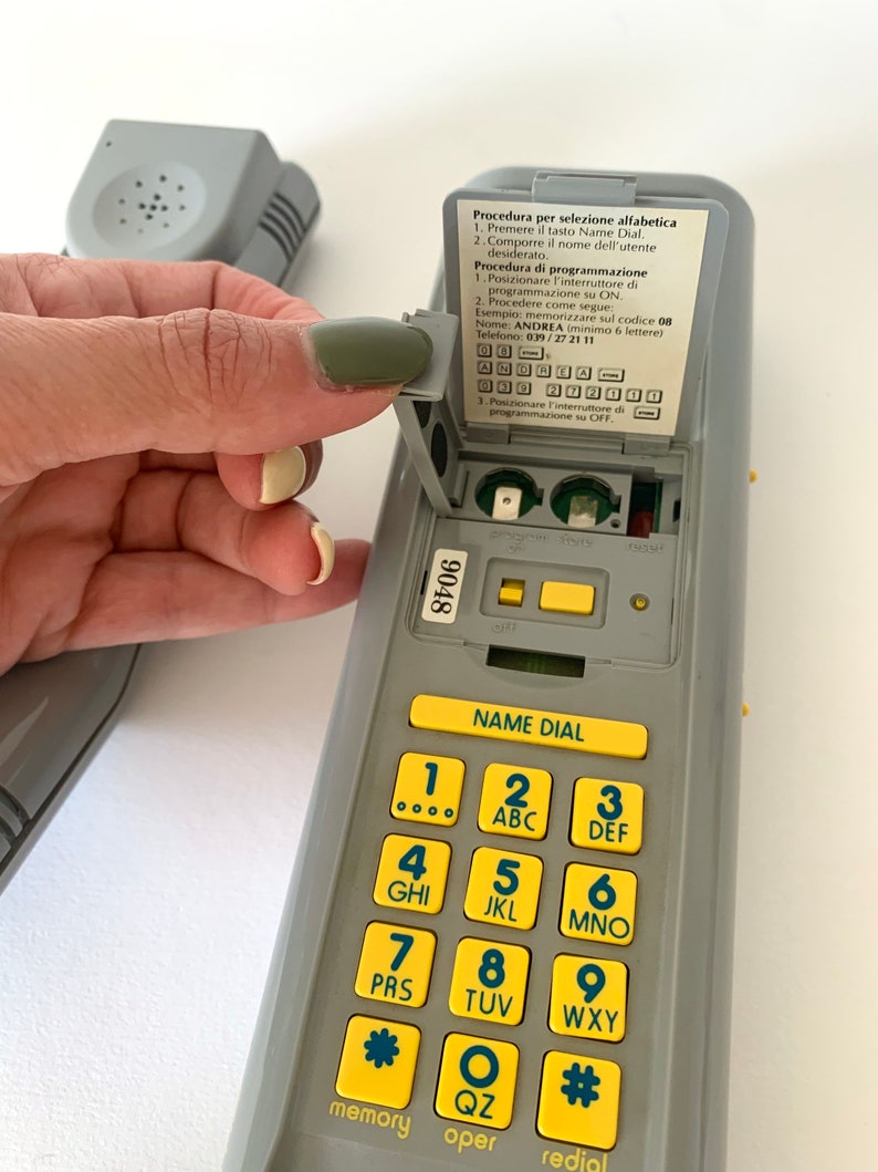 May include: A gray phone with a yellow keypad and a small gray flip-up cover. The cover has instructions for programming the phone in Italian. The keypad has numbers 1 through 9, a star, a circle with the letter "O" inside, and a pound sign.