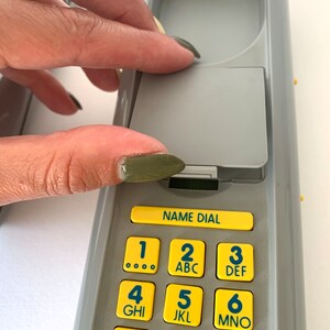May include: A gray phone with a yellow keypad. The keypad has numbers 1 through 9 with letters corresponding to each number. The phone has a name dial button and a gray cover.