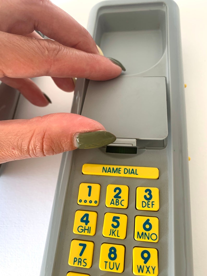 May include: A gray phone with a yellow keypad. The keypad has numbers 1 through 9 with letters corresponding to each number. The phone has a name dial button and a gray cover.