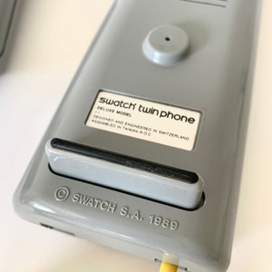 May include: A gray Swatch twin phone with a black cord. The phone has a label that reads "Swatch twin phone Deluxe Model Designed and Engineered in Switzerland Assembled in Taiwan R.O.C." and "&copy; SWATCH S.A. 1989".