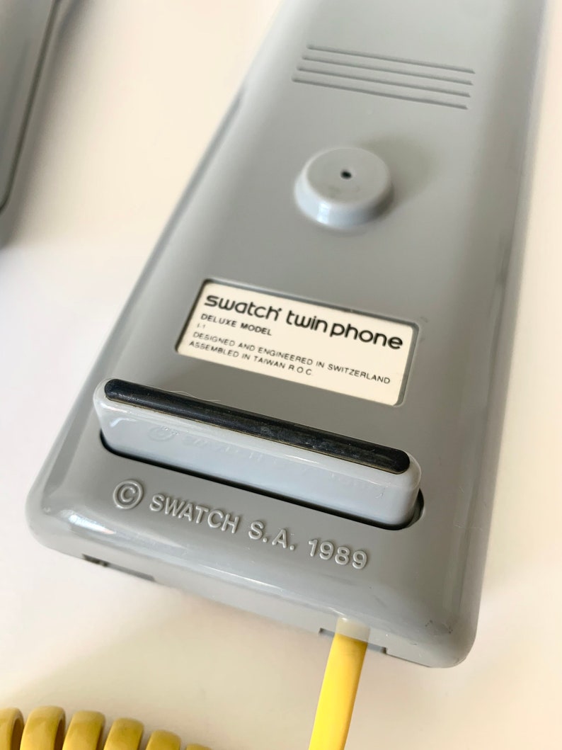 May include: A gray Swatch twin phone with a black cord. The phone has a label that reads "Swatch twin phone Deluxe Model Designed and Engineered in Switzerland Assembled in Taiwan R.O.C." and "&copy; SWATCH S.A. 1989".