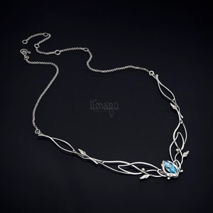 May include: A silver necklace with an intricate, flowing design. The necklace features a central blue gemstone and delicate leaf and vine accents. The chain is thin and elegant, with the brand name "ilmapu" visible on the right side.