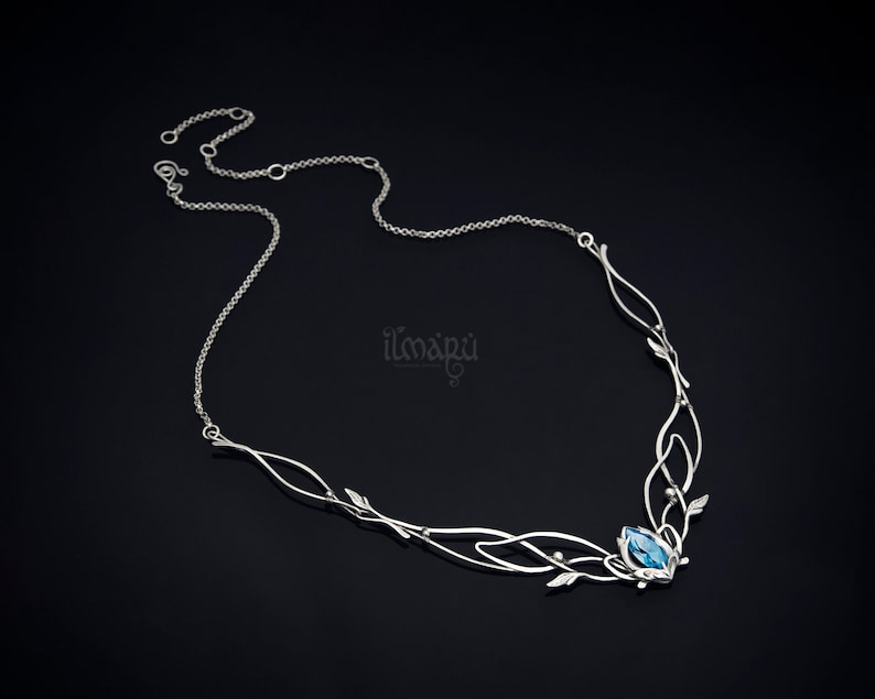 May include: A silver necklace with an intricate, flowing design. The necklace features a central blue gemstone and delicate leaf and vine accents. The chain is thin and elegant, with the brand name "ilmapu" visible on the right side.